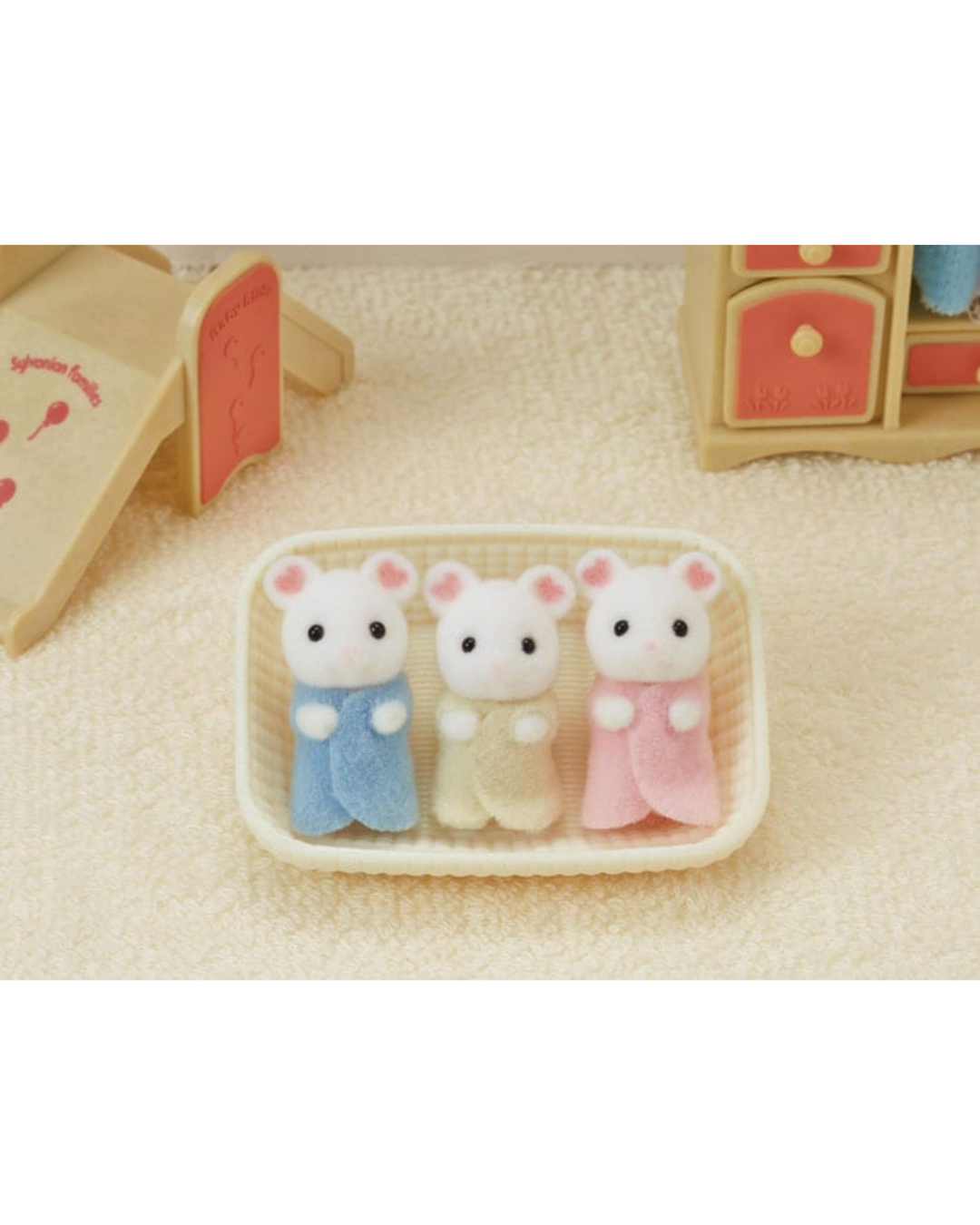 Three small Sylvanian Families marshmallow mice in pastel blue, cream, and pink outfits, arranged in a cream-colored basket on a textured beige surface.