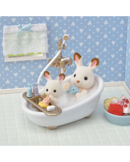 A miniature white bathtub scene with two small white bunny sylvanian figurines sitting in bubble bath, one holding a blue toy and the other resting beside it. The tub has a gold faucet and a small side tray with tiny bath accessories.