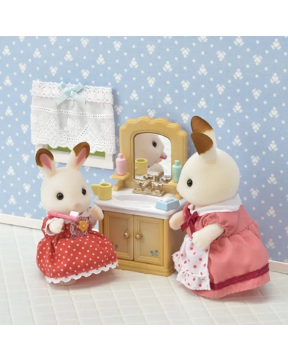 A Sylvanian Bunny  scene featuring two bunny figures in pink and red dresses standing beside a small vanity table with a mirror, set against a blue patterned wallpaper background.