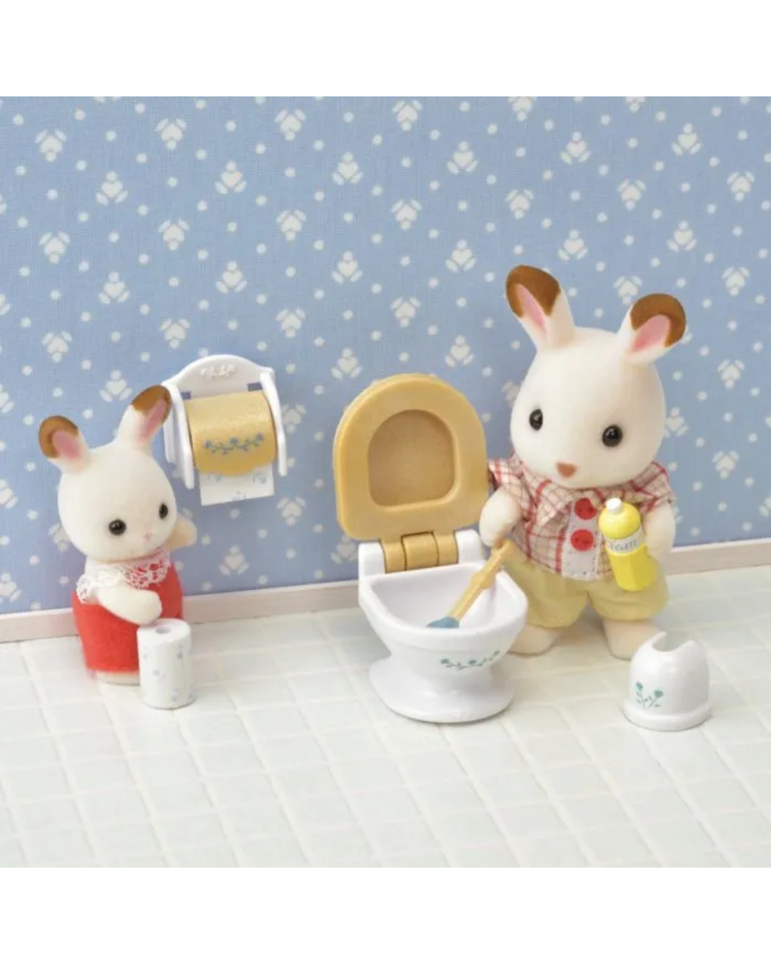 A small sylvanian rabbit figurine wearing a red-and-white checkered shirt and yellow pants, standing in a bathroom and holding a yellow spray bottle next to a white toilet. A second rabbit figurine sits on the left holding a red cup and toilet paper, with matching bathroom accessories and a blue patterned wall background.