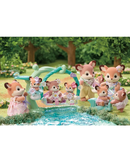 Four sylvanian animal toy figures stand in a sunlit forest setting with trees, grass, and flowers and a lake. The figures are dressed in pastel clothing.