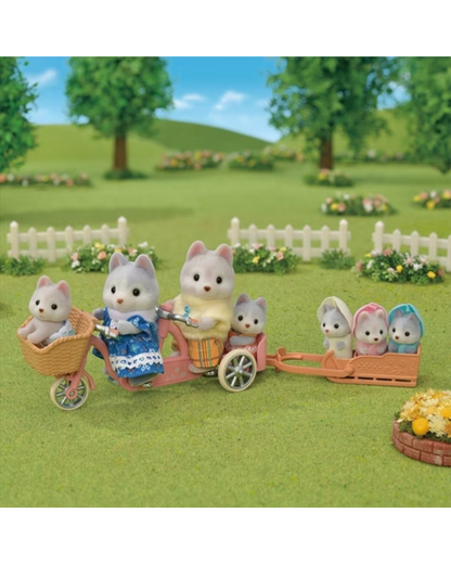 A small sylvanian family Huskey figurine set featuring children and parents in a garden scene. The family rides a pink tricycle with a basket and a wagon attached, set on a green lawn with trees, flowers, and a white picket fence in the background.