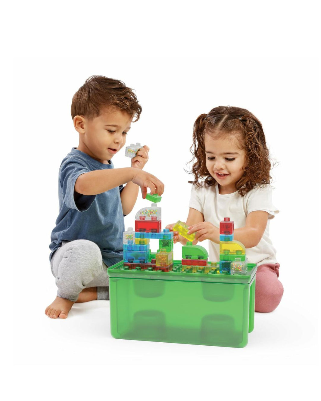 Two children playing with colorful building blocks arranged in a translucent green storage bin on a white studio background.