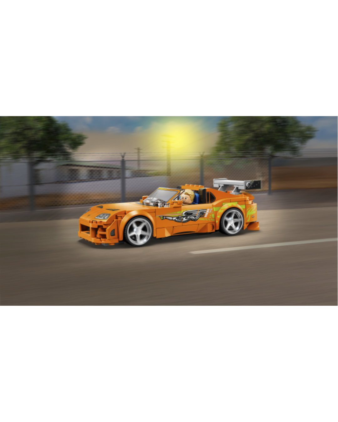 An orange toy sports car built from LEGO, featuring a small minifigure driver, silver wheels, and green and yellow graphic decals, shown against a street background