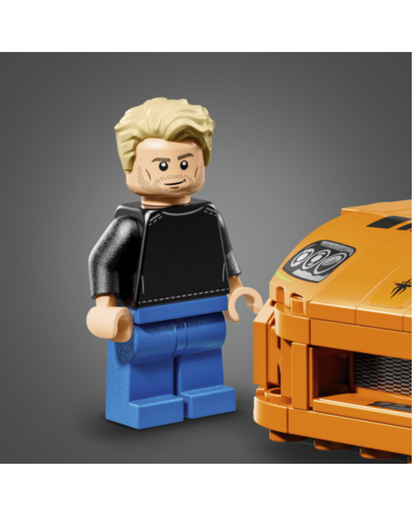 A LEGO minifigure with blond hair, a black shirt, and blue pants standing next to an orange toy car on a gray background.