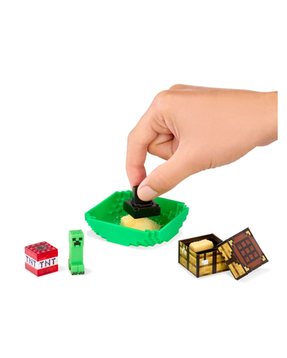 A hand is shown interacting with a Minecraft-themed toy set on a white background. The set includes a green container with a black and yellow piece, a red TNT block, a green Creeper figure, and an open treasure chest.