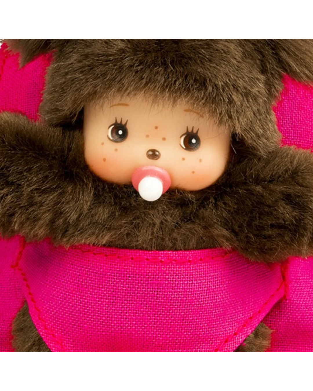 Close-up of Monchhichi with a brown furry hood, peach-colored face, freckles, large dark eyes, a pink pacifier, and a bright pink fabric body.