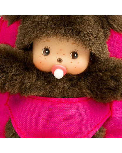Close-up of Monchhichi with a brown furry hood, peach-colored face, freckles, large dark eyes, a pink pacifier, and a bright pink fabric body.
