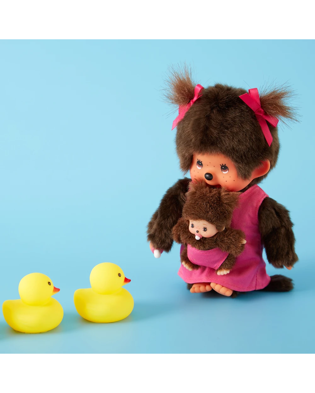 A small brown Monchhichi doll with pink bows in its hair, wearing a pink dress and holding a smaller plush monkey, posed beside two yellow rubber ducks on a light blue background.