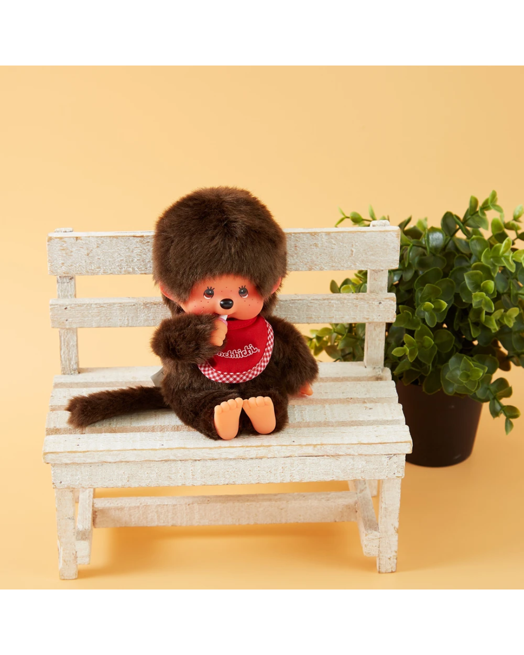 A small brown plush monchhichi doll with peach-colored feet and hands, wearing a red bandana, sitting on a miniature white wooden bench next to a potted green plant against a warm beige background.
