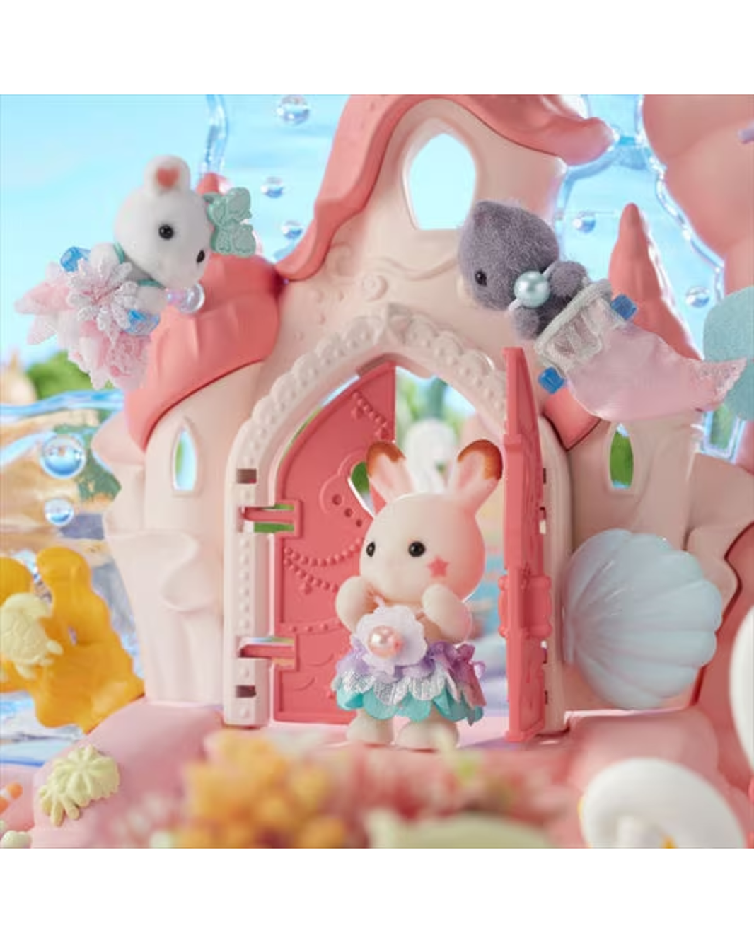 A pastel pink toy castle playset with a small bunny figurine standing in the doorway, plus small animal and shell accessories around it, photographed against a soft blue and pink themed background.