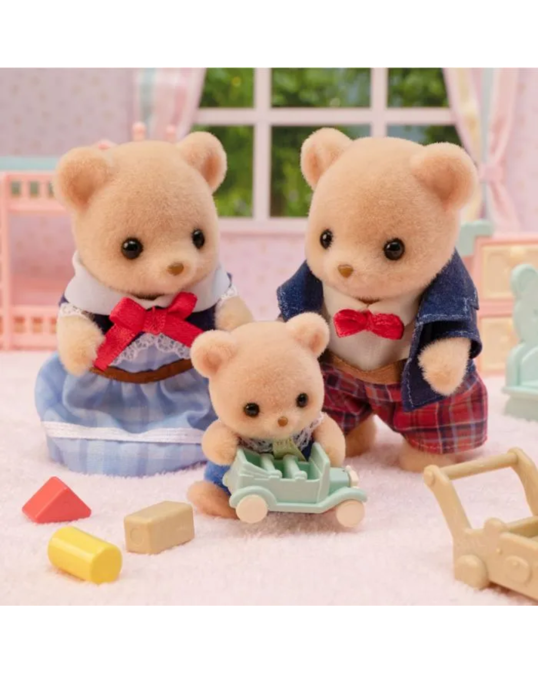 Three Sylvanian Families toy teddy bear characters posed in a dollhouse-style room with miniature blocks and toys. The family set includes a mother bear in a blue dress with a red bow, a father bear in a blue jacket and red plaid shorts with a red bow tie, and a baby bear sitting in a light blue stroller.