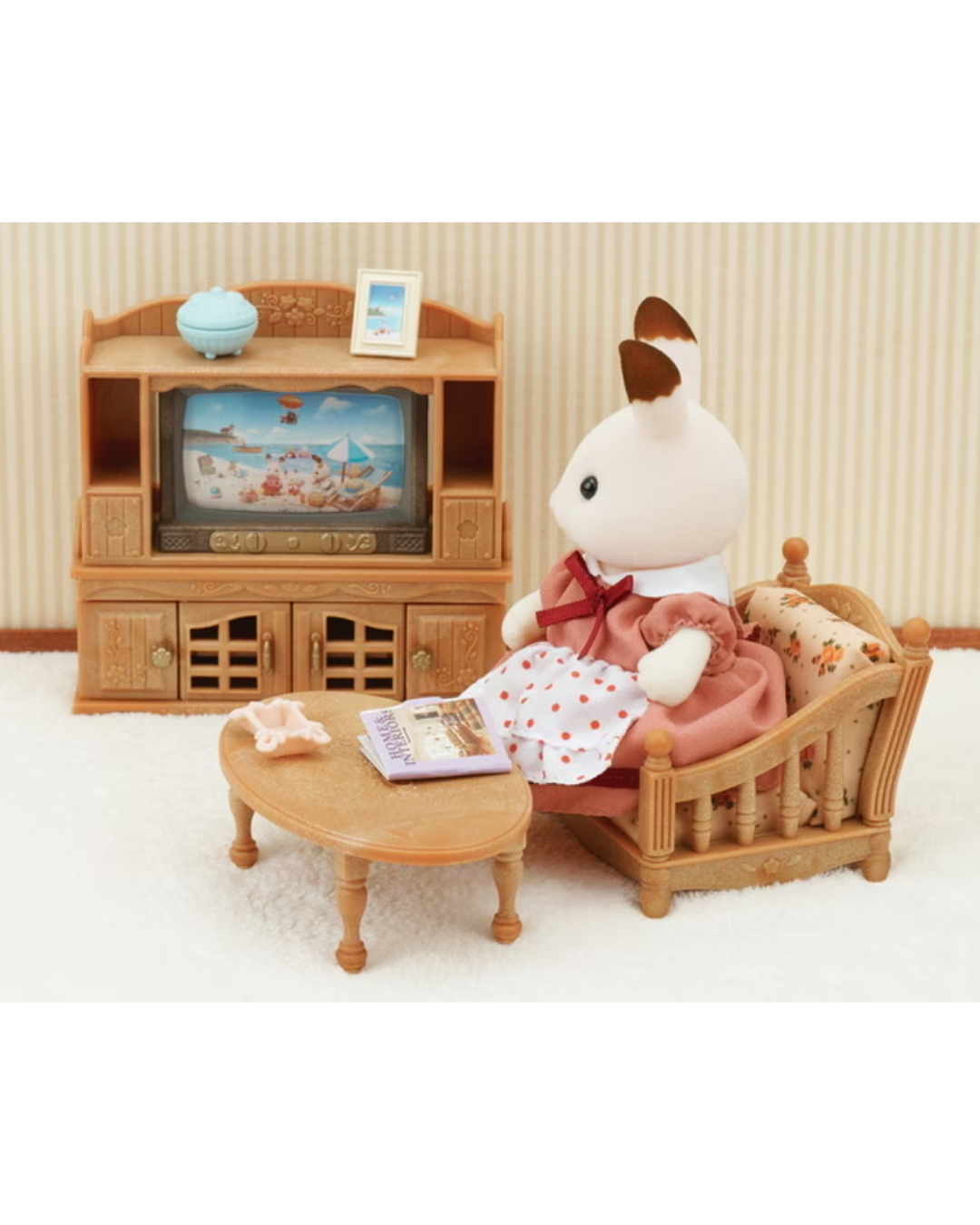 A small Sylvanian Families scene with a white rabbit figure sitting in a wooden armchair next to a round coffee table, with a wooden TV cabinet in the background.