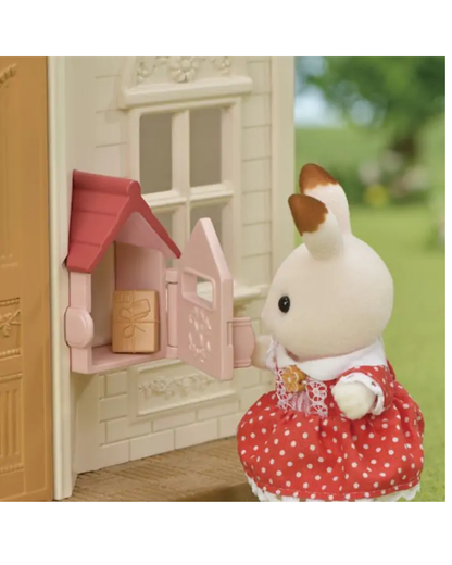 A Sylvanian Families toy figure of a rabbit in a red and white polka dot dress stands next to a miniature pink and cream house with a small red-roofed window display.