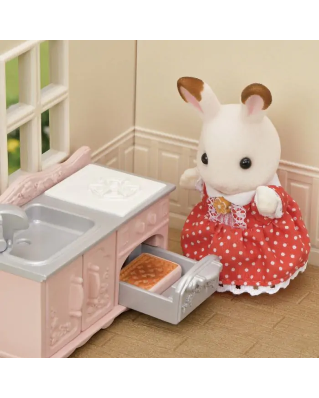 A small white rabbit toy wearing a red dress with white polka dots stands beside a pink miniature kitchen cabinet with a sink and an open oven containing a waffle, set against a beige room backdrop.
