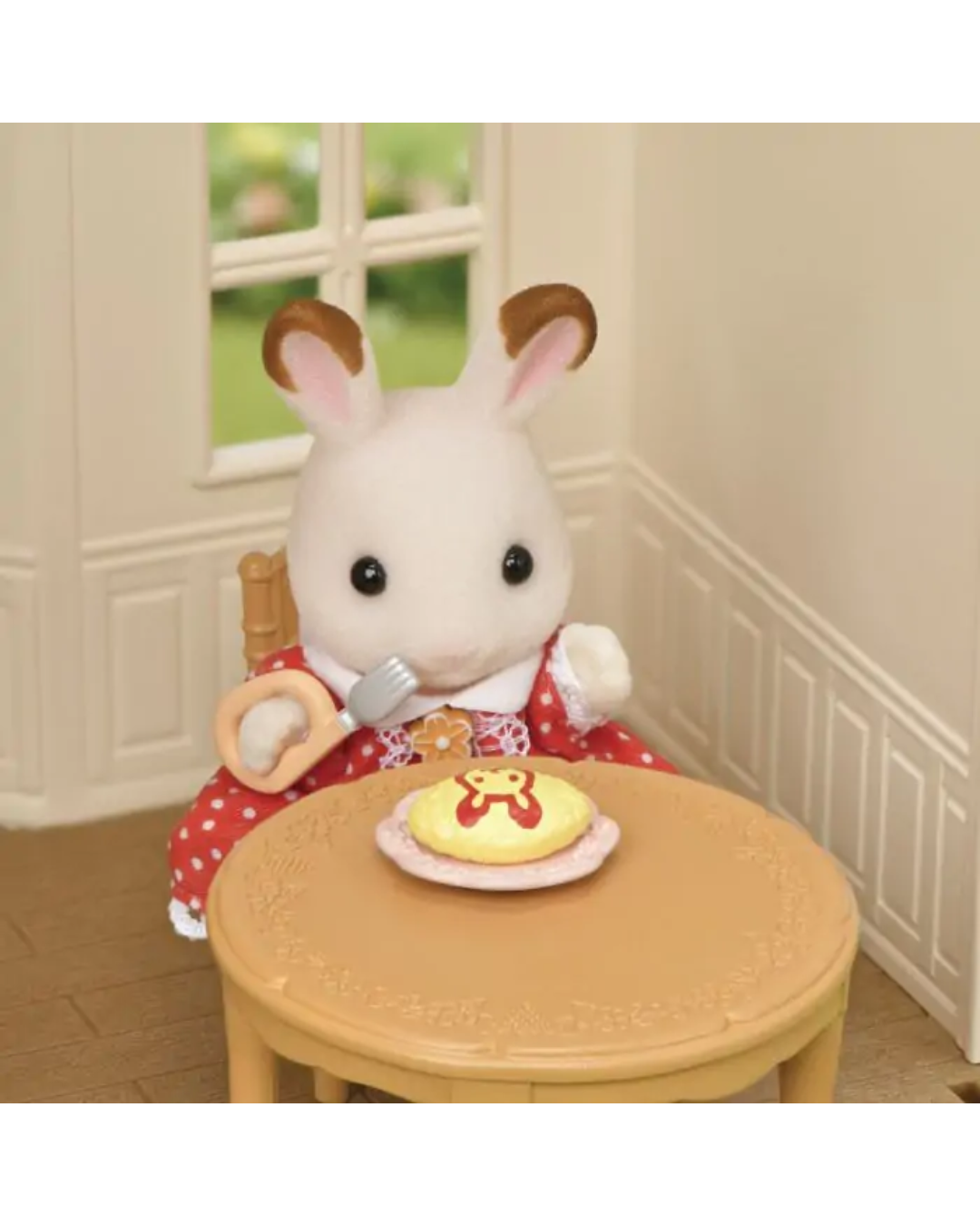 A small white rabbit sylvanian character wearing a red dress with white polka dots sits at a round wooden table, holding a fork, with a gingerbread cookie on a plate in front of it.