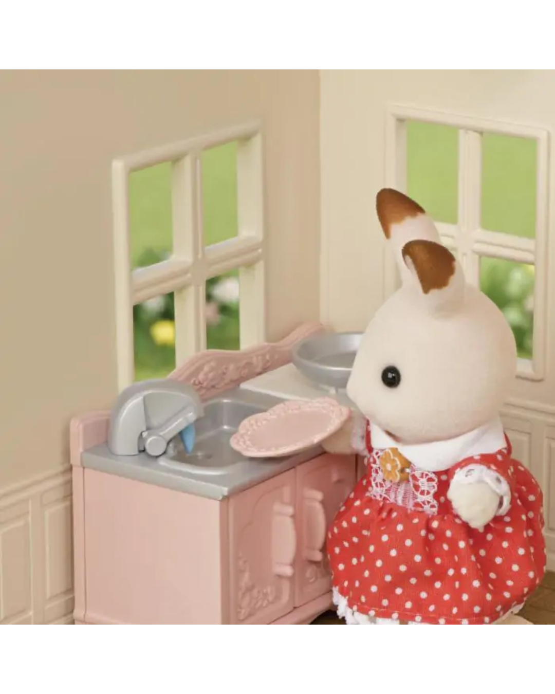 A small rabbit toy figure in a red polka-dot dress stands beside a pink dollhouse kitchen sink with a plate and faucet.