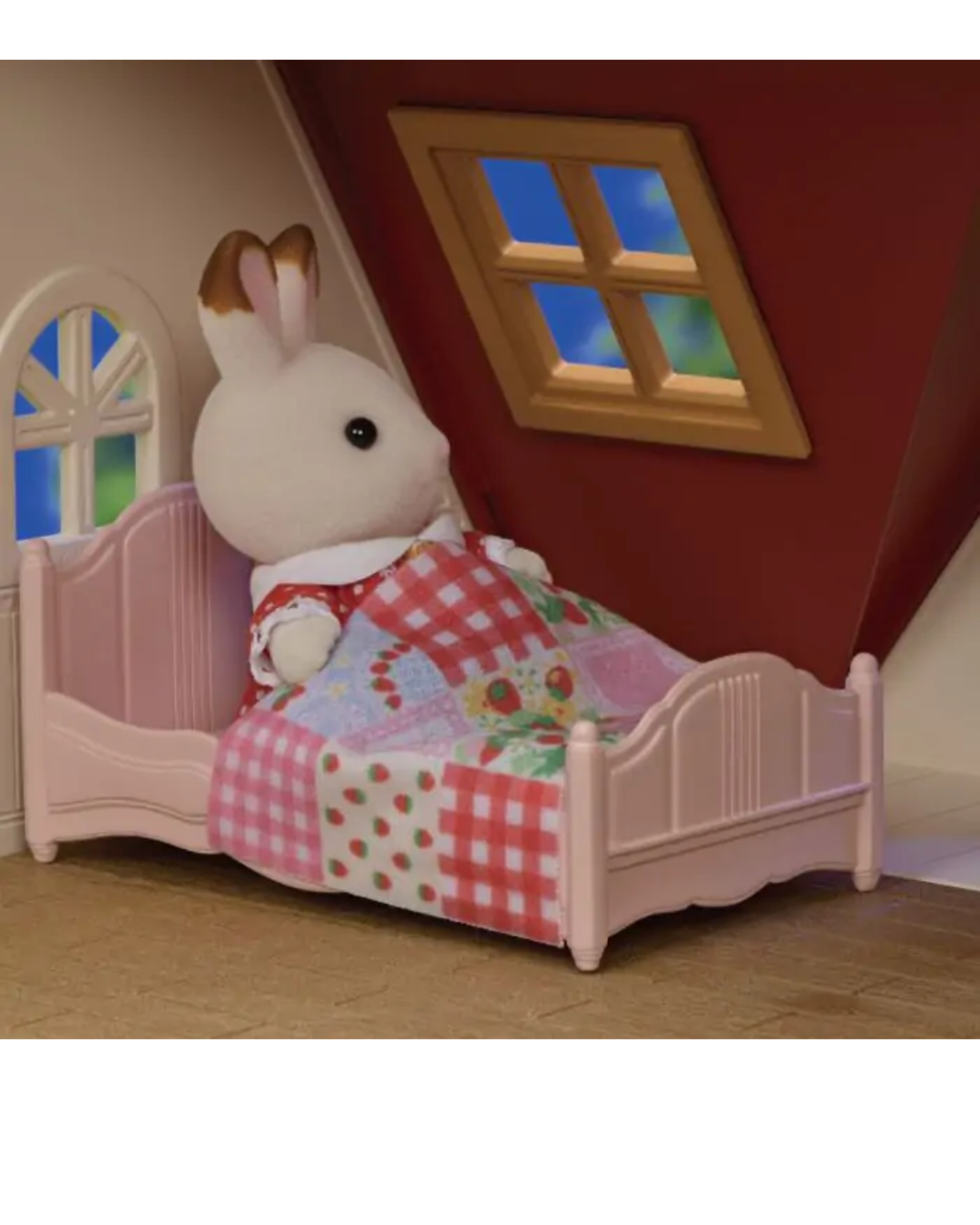 A small white rabbit plush toy with brown-tipped ears sits upright in a pink wooden bed frame, covered with a red-and-white gingham blanket and a multicolor floral quilt, inside a dollhouse-like room with a red wall and window.
