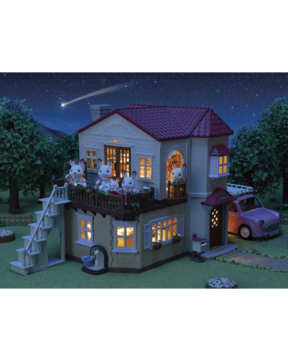 A Sylvanian Families model of a two-story house at night, featuring illuminated windows, a balcony with rabbit characters, an external staircase, a small car in a driveway, and a starry sky with a shooting star in the background.