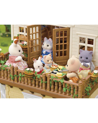 A Sylvanian Families miniature dollhouse scene showing several animal characters gathered on a balcony having a tea party. The house has cream-colored siding, brown wooden balcony railings and doors, and green ivy details.