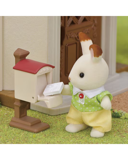 A Sylvanian Families toy figure of a rabbit character in a green and yellow outfit standing next to a small mailbox on a grassy surface.