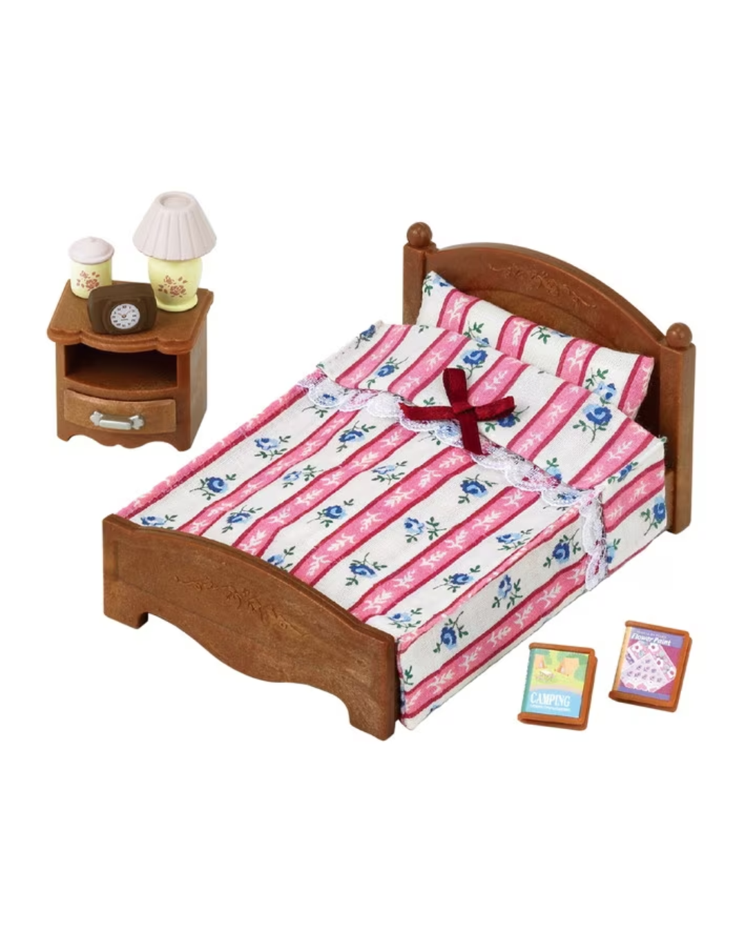 A Sylvanian Families bed with a wooden frame, pink and white striped bedding with blue floral accents, a matching nightstand, a small lamp, and two small accessory boxes on a plain white background.