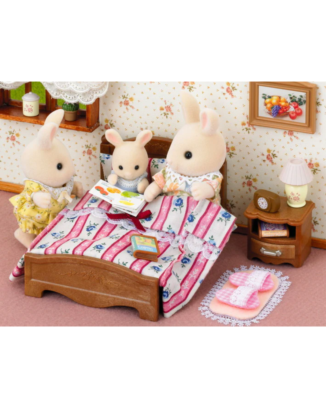 A Sylvanian Families bedroom scene with three rabbit toy figures around a small bed. One rabbit sits on the bed reading a book, while another stands on the left in a yellow dress and the third sits on the right in a cream outfit.