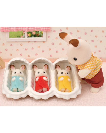 Three Sylvanian Families plush baby rabbits in white cribs, each wearing a different colored blanket (blue, red, yellow), with a small plush rabbit figure in a yellow-and-white check shirt and red pants standing beside them against a pink floral background.