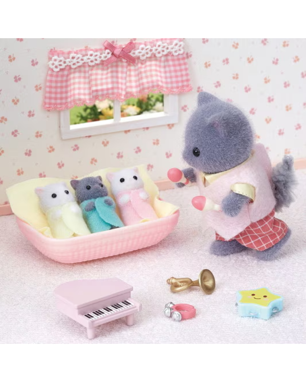 A miniature dollhouse-style nursery scene with a gray cat-like toy figure standing beside a pink bassinet holding pink balls, with three small plush toys inside the bassinet. A pink toy piano, brass instrument, ring, and star-shaped accessory are arranged on a light pink floor against a floral wallpaper background.