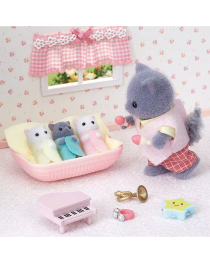 A miniature dollhouse-style nursery scene with a gray cat-like toy figure standing beside a pink bassinet holding pink balls, with three small plush toys inside the bassinet. A pink toy piano, brass instrument, ring, and star-shaped accessory are arranged on a light pink floor against a floral wallpaper background.
