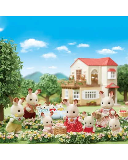 A family of Sylvanian rabbits gathered outdoors in a flowerbed with a small house and trees in the background. The rabbits are dressed in colorful clothing and appear to be playing or having a picnic in a staged garden scene.