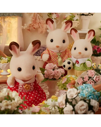 Three Sylvanian Family rabbit toys posed together in a floral, warmly lit indoor scene. The rabbits wear colorful outfits, with pink, red, green, and white clothing, and are surrounded by artificial flowers and a small soccer ball.