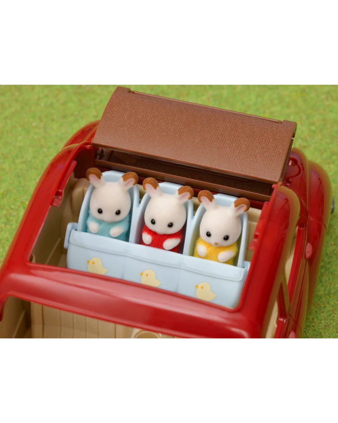 A close-up of a Sylvanian Families vehicle with the back open, showing three small animal figurines sitting inside a blue stroller. The background is a green textured surface.